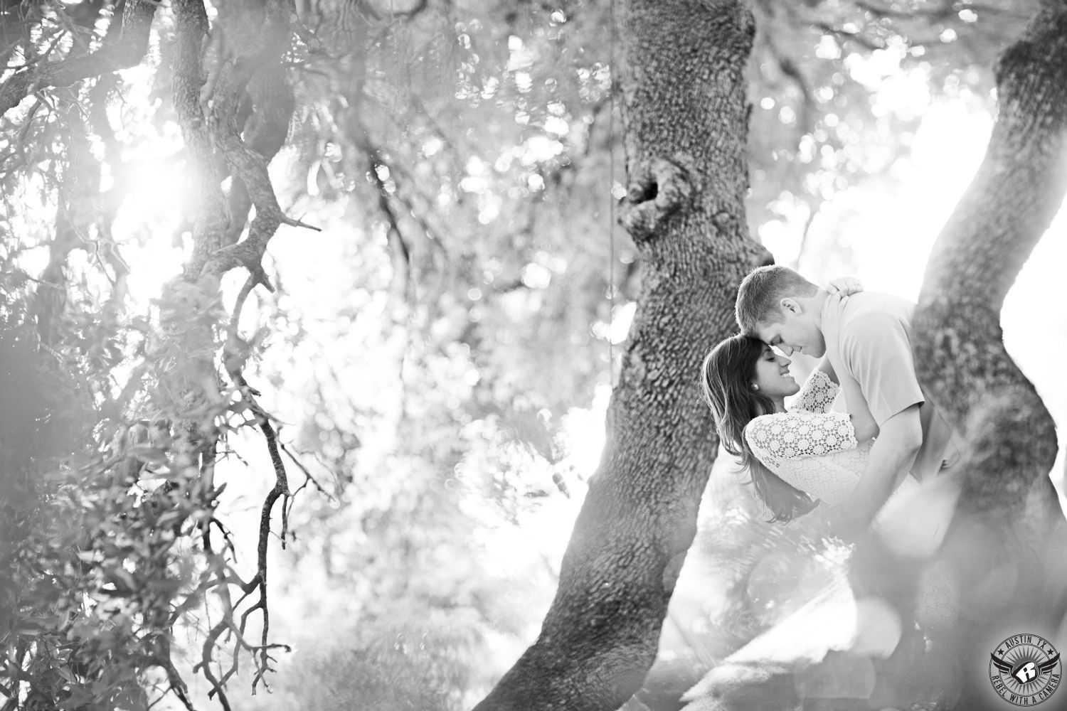 Long haired brunette girl wearing a short sleeve lace blouse smiles as she holds a sandy haired guy wearing a light colored casual dress shirt surrounded but tree branches with glowing  sun popping through at the Texas State Capital in this serene engagement picture in Austin, Texas. 